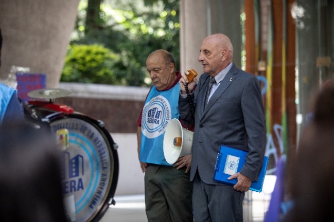 A person in a suit holding a speaker and a person in a blue shirt

Description automatically generated